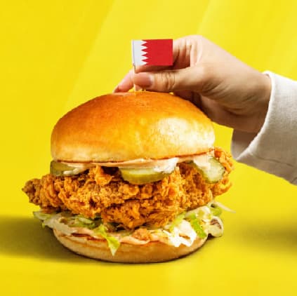A hand placing a Bahrain flag on a Sandwich Guys chicken burger